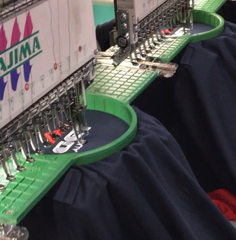 Tajima multi-head embroidery machine stitching a GET logo onto navy shirts held in green hoops